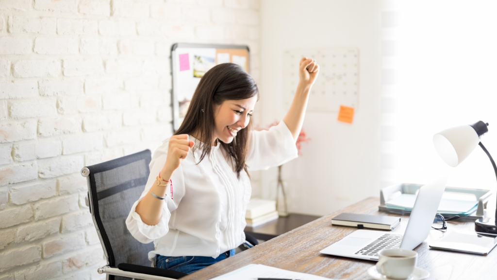A businesswoman at a desk, raising one fist in excitement, in an open and airy office.