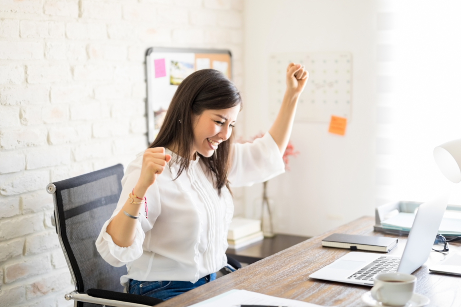 A businesswoman at a desk, raising one fist in excitement, in an open and airy office.