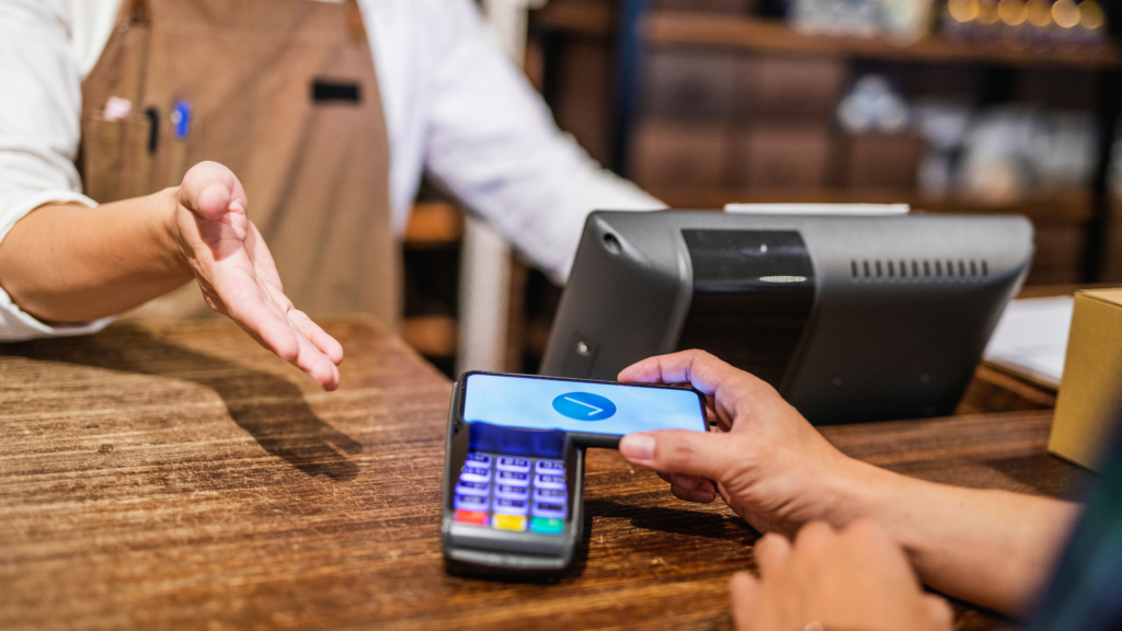 A cashier and customer exchanging a credit card across a counter, with the cashier wearing an apron and standing in front of a POS while the customer holds their card over a card reader.