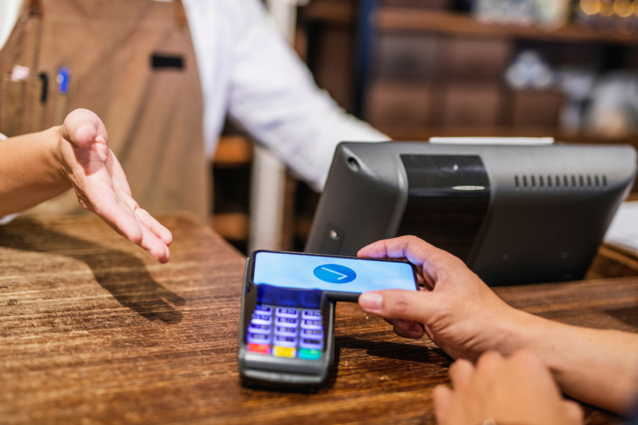 A cashier and customer exchanging a credit card across a counter, with the cashier wearing an apron and standing in front of a POS while the customer holds their card over a card reader.