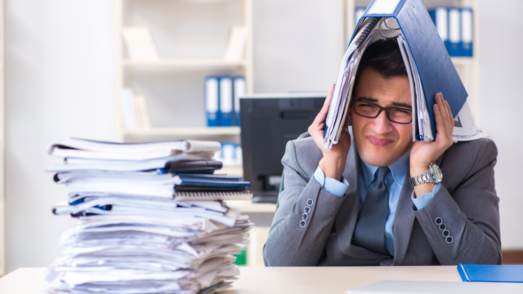 A frustrated man sitting at a desk, holding a book over his head, surrounded by a stack of books.