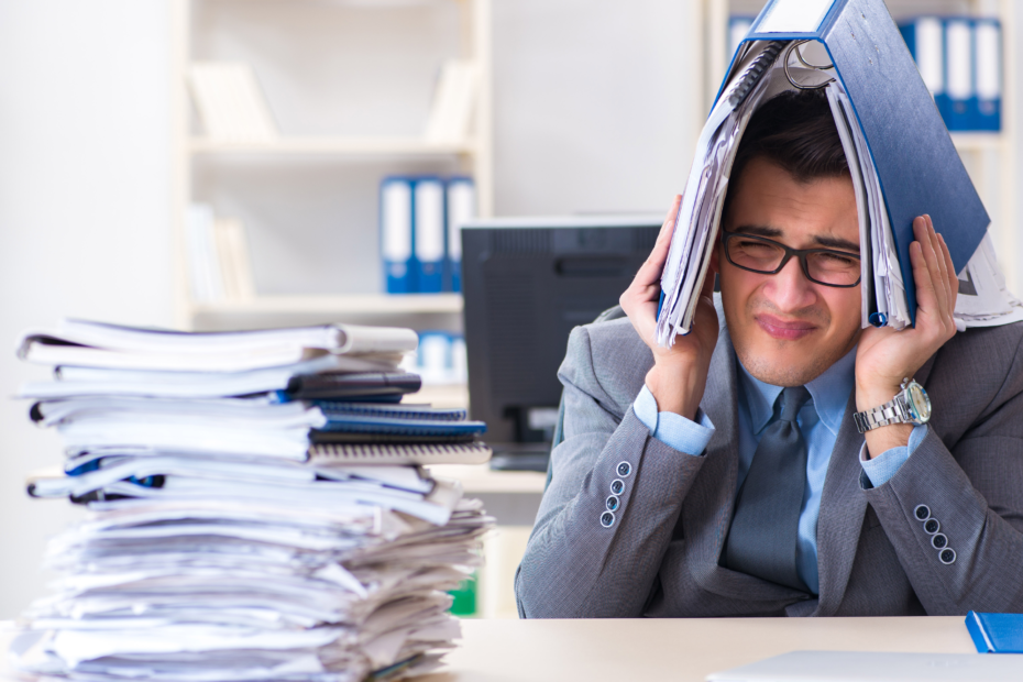 A frustrated man sitting at a desk, holding a book over his head, surrounded by a stack of books.