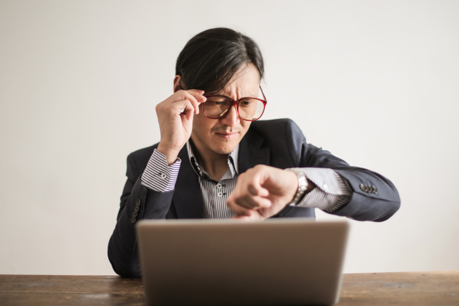 A man in business attire, sitting at a desk, lowering his glasses to look at his watch.