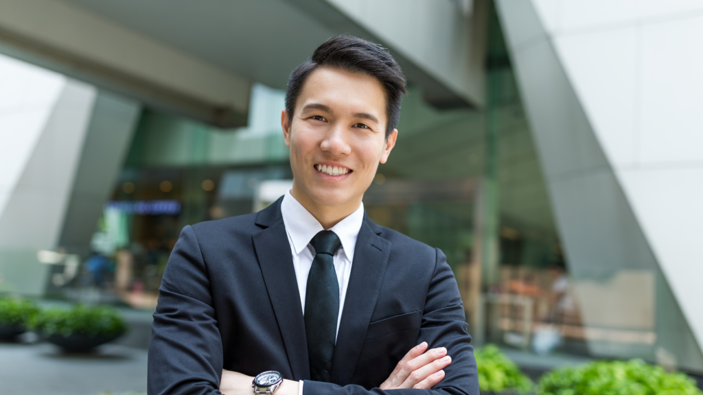 A smiling businessman with arms crossed, standing in front of a modern glass business building.