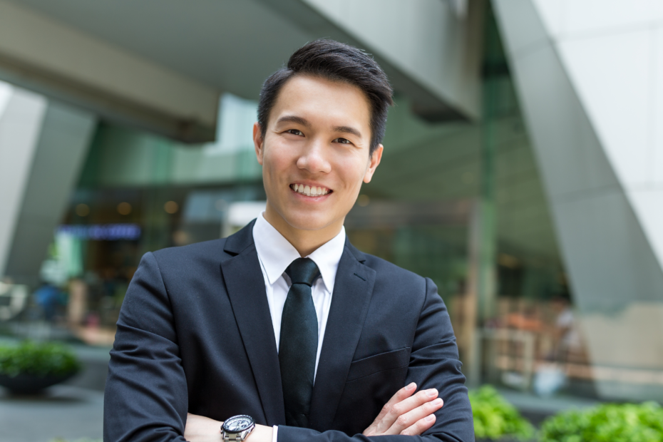 A smiling businessman with arms crossed, standing in front of a modern glass business building.