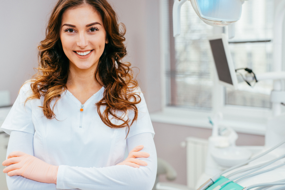 A young female dentist with a warm smile and crossed arms, dressed in a white lab coat, standing confidently in a dentist's office.