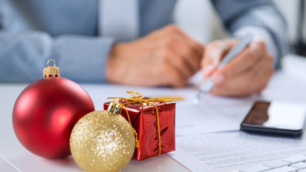 Business person's arms writing with a pencil at a desk, with red and gold holiday ornaments in the foreground.