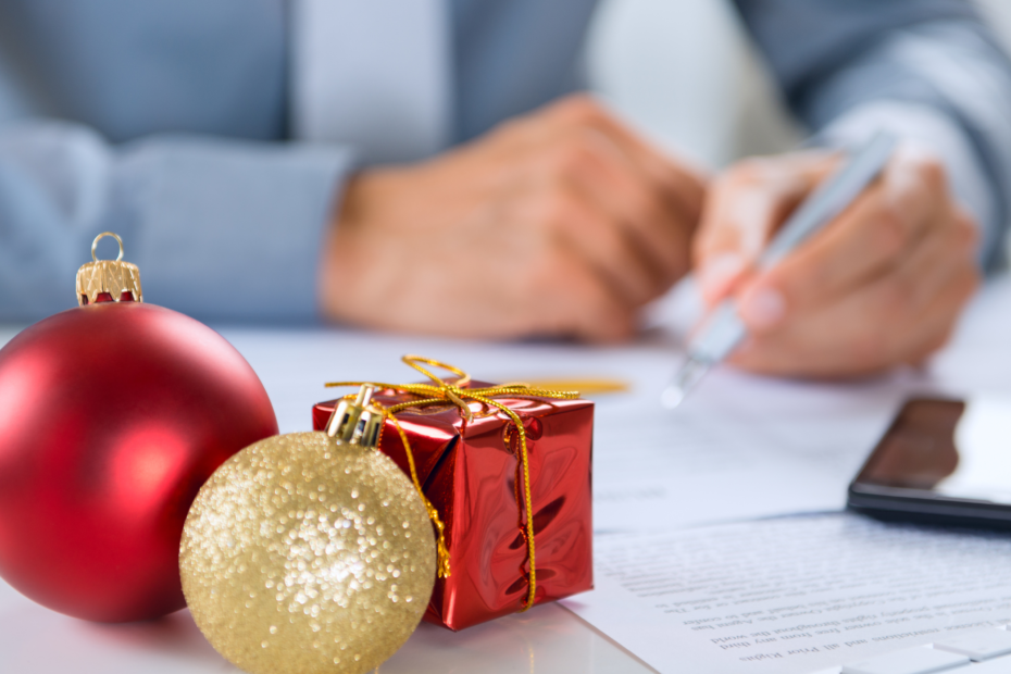 Business person's arms writing with a pencil at a desk, with red and gold holiday ornaments in the foreground.