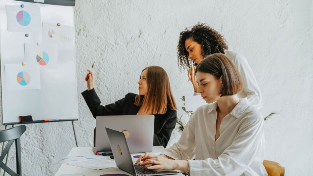 Three diverse women focused on a whiteboard with graphs in an office.