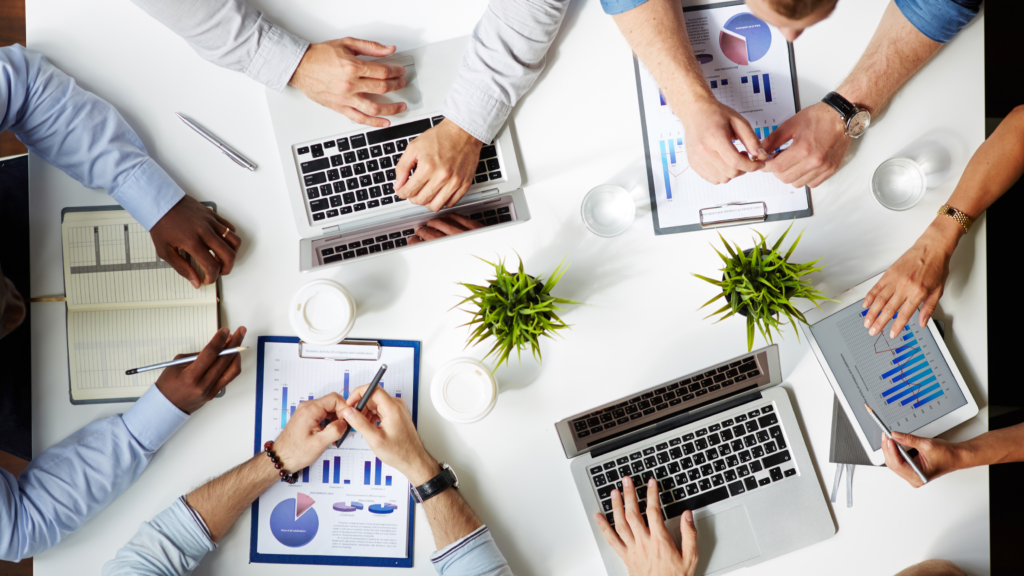 Top-down view of six pairs of hands working on computers in a diverse office setting.