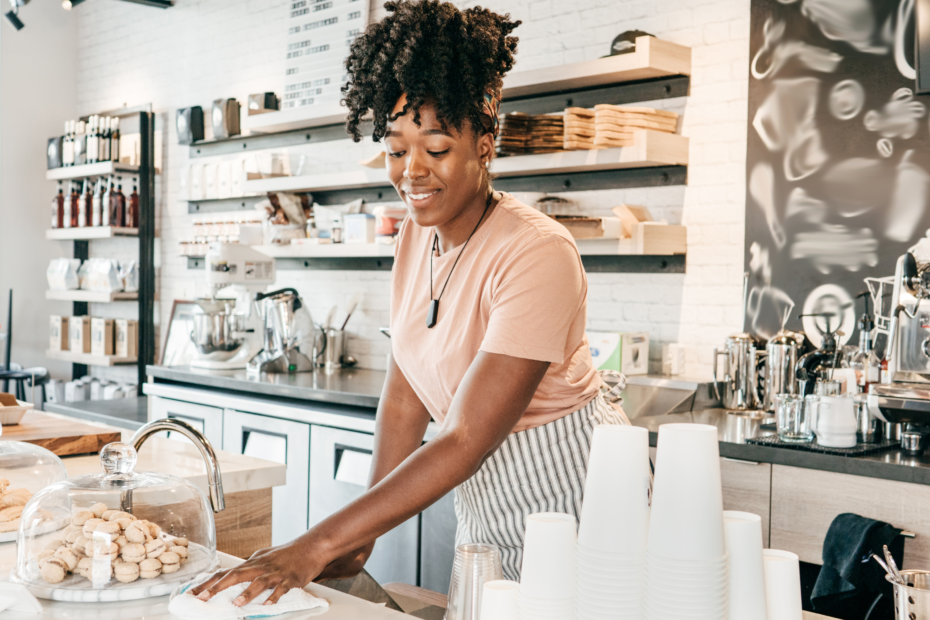 Woman of color behind the counter of a light and airy bakery coffee shop surrounded by pastries and an espresso machine.