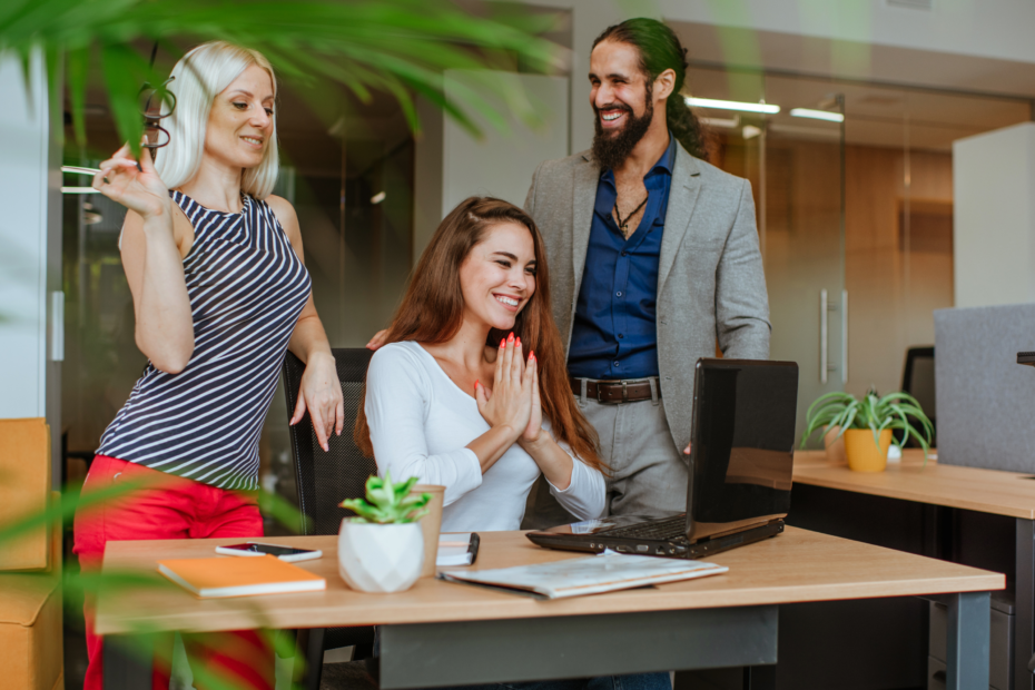 Three people, two females and one male, smiling in an office with plants. One female seated at a desk.