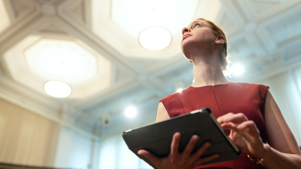 Businesswoman holding a tablet and looking confident in a room with elaborate ceiling tiles.