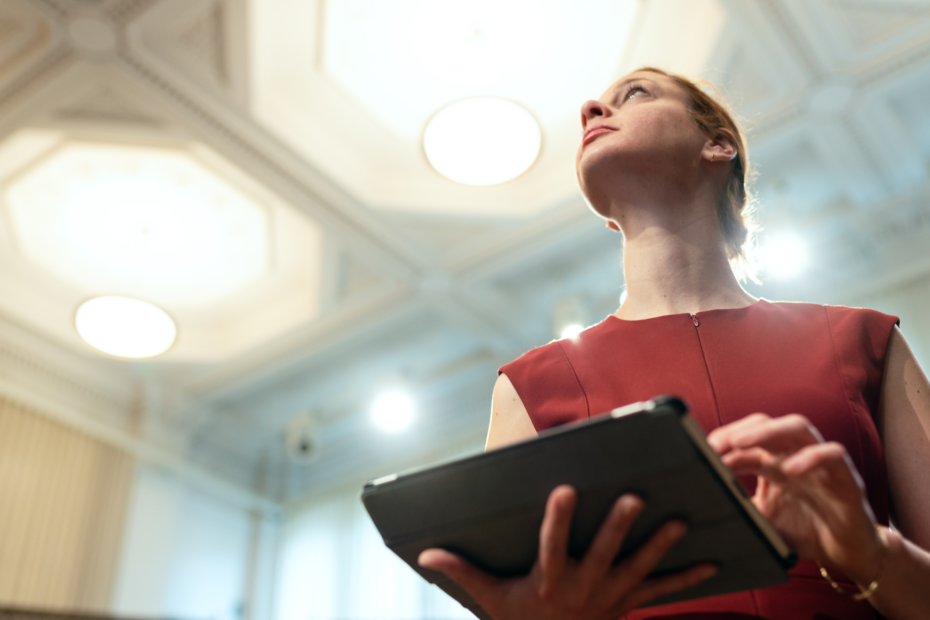 Businesswoman holding a tablet and looking confident in a room with elaborate ceiling tiles.