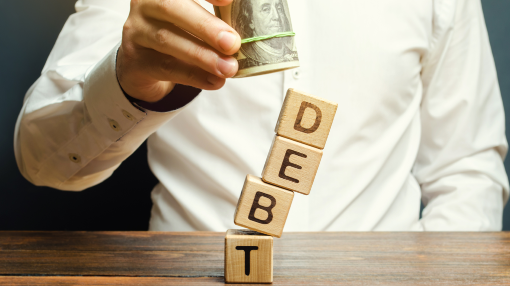 A man's hands carefully stack wooden blocks that spell out the word "DEBT," while holding a bundle of dollar bills in the background.
