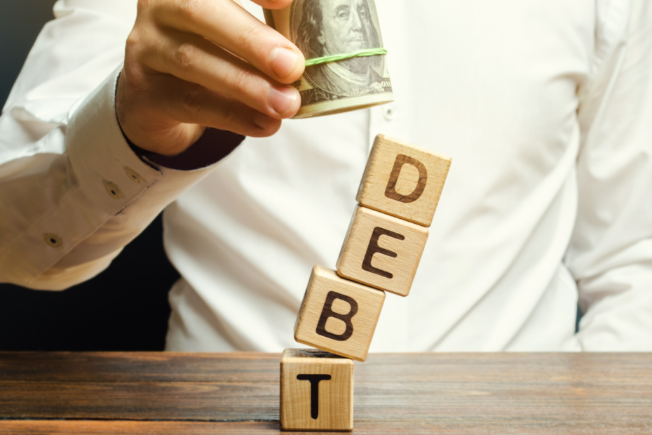 A man's hands carefully stack wooden blocks that spell out the word "DEBT," while holding a bundle of dollar bills in the background.