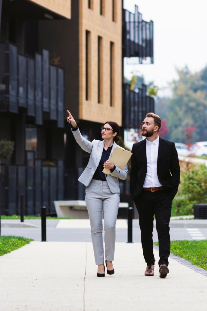 A property manager and a client surveying a property in a modern neighborhood with a very edgy building design behind them as they walk down a sidewalk.